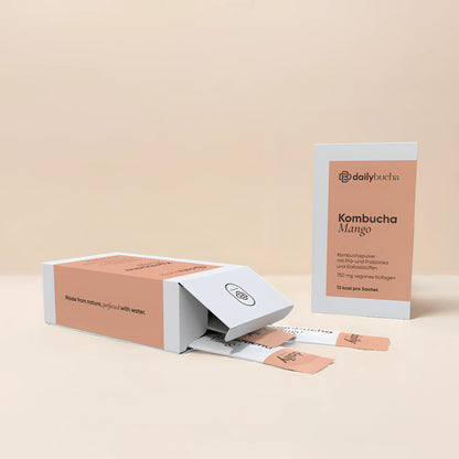 Daily Bucha Mango-Kombucha mit veganem Kollagen und Ballaststoffen, ohne Zuckerzusatz – geöffnete Verpackung mit Sachets und aufgestellter Box vor beigem Hintergrund